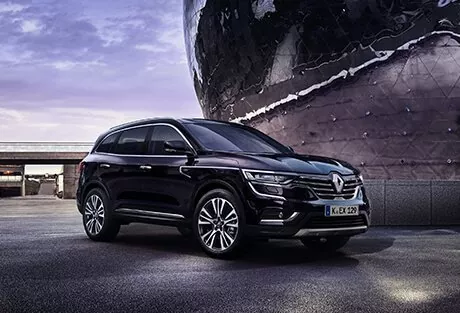 Ein schwarzer Renault-Geländewagen parkt in der Abenddämmerung auf einer gepflasterten Fläche in der Nähe eines modernen, reflektierenden Gebäudes, mit einem violetten Himmel im Hintergrund.