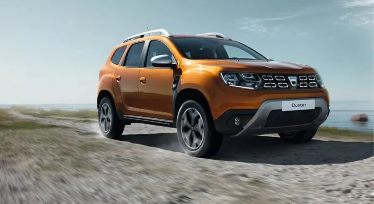 Ein orangefarbener Dacia Duster SUV fährt auf einer Schotterstraße in der Nähe eines Gewässers, mit Gras und Felsen an den Seiten und einem klaren Himmel im Hintergrund. Der abenteuerliche Dacia Duster hebt sich von der malerischen Landschaft ab.