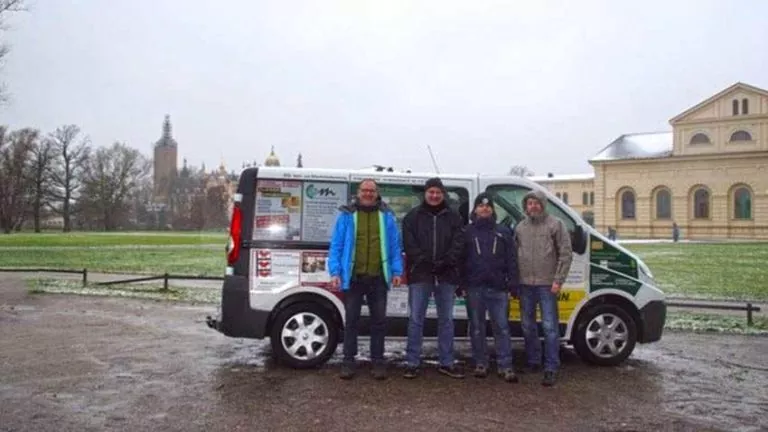 Vier Personen in Winterkleidung stehen vor einem Lieferwagen, der mit verschiedenen Plakaten und Schildern beklebt ist. Im Hintergrund sind historische Gebäude und ein verschneiter Park zu sehen.