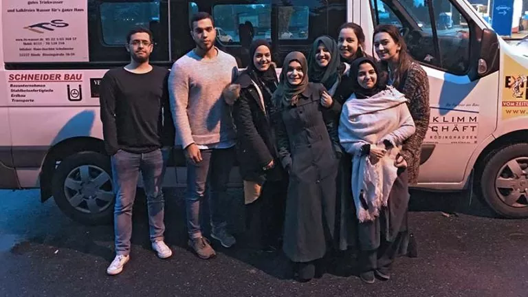 Eine Gruppe von acht jungen Erwachsenen, von denen einige einen Hidschab tragen, steht in der Abenddämmerung lächelnd vor einem geparkten Lieferwagen auf einer nassen Straße.