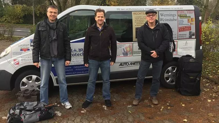 Drei Männer stehen vor einem mit Werbung bedeckten Lieferwagen, jeder mit einer Reisetasche zu Füßen. Die Szene spielt sich im Freien ab, mit Herbstblättern auf dem Boden und Bäumen im Hintergrund.