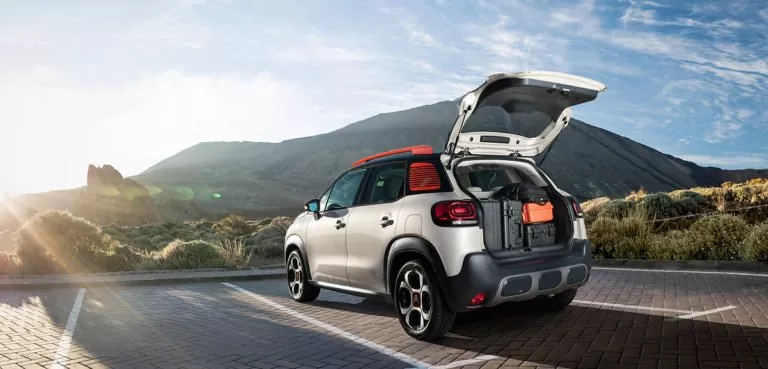 Ein silberner C3 Aircross Geländewagen mit orangefarbenem Dach und offenem Kofferraum ist auf einem leeren Parkplatz geparkt, das Gepäck liegt darin. Berge und Sonnenlicht bilden eine malerische Kulisse.