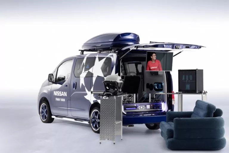 Ein blauer Nissan NV200 Evalia Fan Van mit geöffneter Heckklappe zeigt einen Fernseher, Spielgeräte und einen Sitzbereich. Im Inneren sitzt eine Frau in einem roten Hemd. Ein blauer Sessel draußen neben dem Van schafft einen gemütlichen Unterhaltungsraum.
