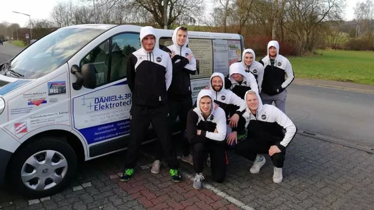 Acht Männer in passenden schwarz-weißen Trainingsanzügen posieren und lächeln vor einem Lieferwagen mit Geschäftswerbung an der Seite, der auf einer Straße mit Bäumen und Gras im Hintergrund geparkt ist.