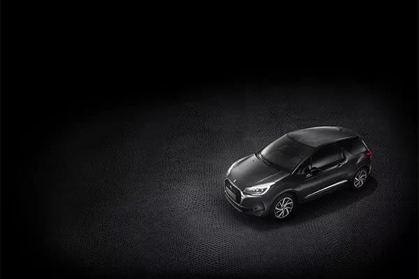 Ein dunkel gefärbtes Auto wird auf einer strukturierten, schwarzen Fläche unter dramatischer Beleuchtung geparkt, wobei der Großteil des Hintergrunds in der Dunkelheit verschwindet.