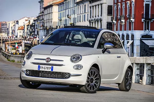 Ein weißer Fiat 500 ist an einem sonnigen Tag auf einer Straße in der Nähe eines Kanals geparkt, mit bunten Gebäuden und einem Geländer im Hintergrund.