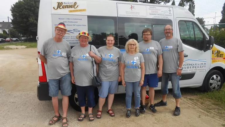 Sechs Personen stehen lächelnd vor einem weißen Lieferwagen mit Werbung und Logos darauf. Sie alle tragen passende blaue Mölln-T-Shirts, einige haben Hüte und Brillen auf. Im Hintergrund sind ein Zaun und Bäume zu sehen.