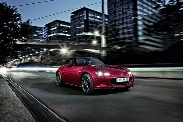 Ein roter Cabrio-Sportwagen fährt in der Dämmerung schnell durch eine Stadtstraße, mit modernen Glasgebäuden und verschwommenen Lichtern im Hintergrund, die Geschwindigkeit und Bewegung betonen.