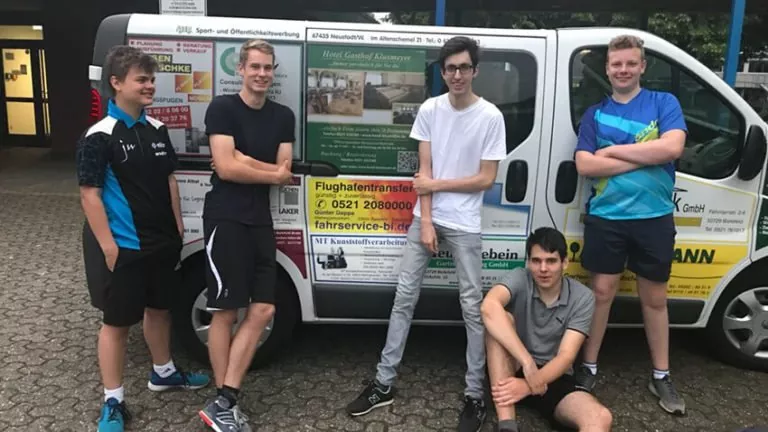 Fünf junge Männer in sportlicher Kleidung posieren und lächeln vor einem Lieferwagen, der mit verschiedenen Geschäftsanzeigen beklebt ist. Eine Person sitzt auf dem Boden, während die anderen um sie herumstehen. Sie wirken alle entspannt und freundlich.