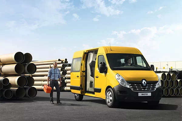 Eine Person geht neben einem gelben Renault Master Transporter, der in der Nähe von großen Stapeln von Betonrohren unter einem teilweise bewölkten Himmel geparkt ist. Die Seitentür des Lieferwagens ist offen, und die Person trägt einen orangefarbenen Container.