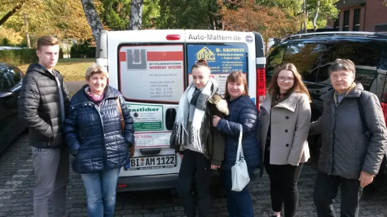 Sechs Personen stehen dicht beieinander vor einem weißen Lieferwagen, auf dessen Rückseite verschiedene Geschäftsreklamen angebracht sind. Im Hintergrund sind Bäume und ein Gebäude zu sehen, und die Gruppe scheint sich an einem sonnigen Tag im Freien aufzuhalten.