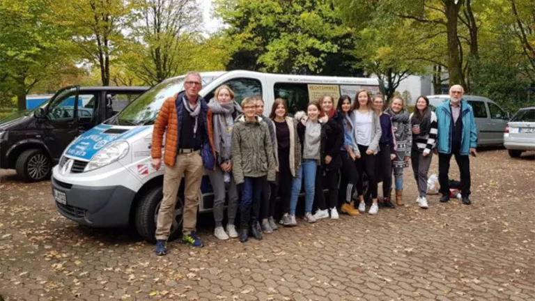 Eine Gruppe von Menschen, darunter Erwachsene und Jugendliche, steht in einer Reihe und lächelt vor einem weißen Lieferwagen, der auf einem Ziegelsteinpflaster geparkt ist, mit Bäumen und parkenden Autos im Hintergrund.