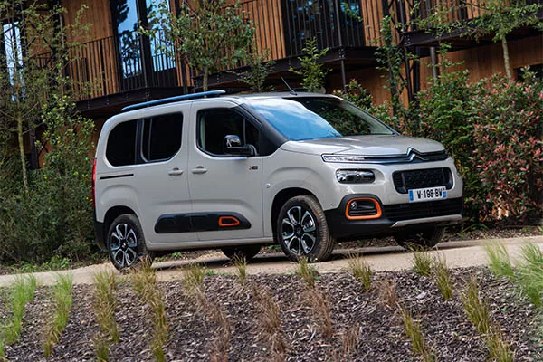 Ein grauer Citroën Berlingo parkt auf einer gepflasterten Einfahrt, umgeben von Grün und Pflanzen, mit einem modernen Holzgebäude im Hintergrund.