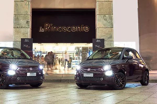 Zwei schwarze Fiat 500 mit eingeschalteten Scheinwerfern parken vor dem beleuchteten Eingang von la Rinascente, einem Kaufhaus. Durch die Glastüren im Hintergrund sind Menschen zu sehen.