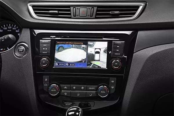 Nahaufnahme des Armaturenbretts mit Rückfahrkamera, Einparkhilfe, Umgebungsansicht und Bedientasten für Audio-, Klima- und Navigationssysteme. Der Bildschirm zeigt eine Warnung an, die Umgebung zu überprüfen, bevor man manövriert.