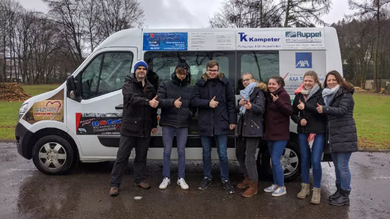 Eine Gruppe von sieben Personen steht an einem regnerischen Tag vor einem weißen Lieferwagen, alle lächeln und zeigen die Daumen nach oben. Der Lieferwagen zeigt verschiedene Firmenlogos und Werbung. Im Hintergrund sind Bäume und Gras zu sehen.