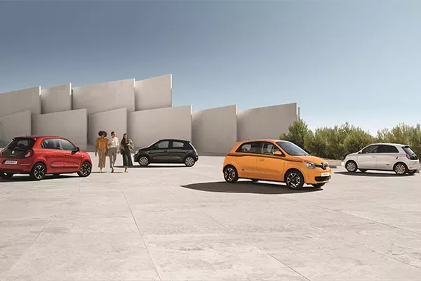 Fünf kleine Autos in verschiedenen Farben parken und fahren auf einer großen gepflasterten Fläche in der Nähe eines modernen Gebäudes mit kantigen Betonwänden. Drei Personen stehen zusammen und unterhalten sich in der Mitte des Bildes. Im Hintergrund sind Bäume zu sehen.