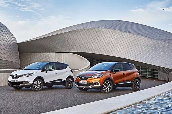Zwei Renault Captur Geländewagen, ein weißer und ein orangefarbener, sind nebeneinander auf einer gepflasterten Fläche vor einem modernen, geschwungenen Metallgebäude unter blauem Himmel geparkt.