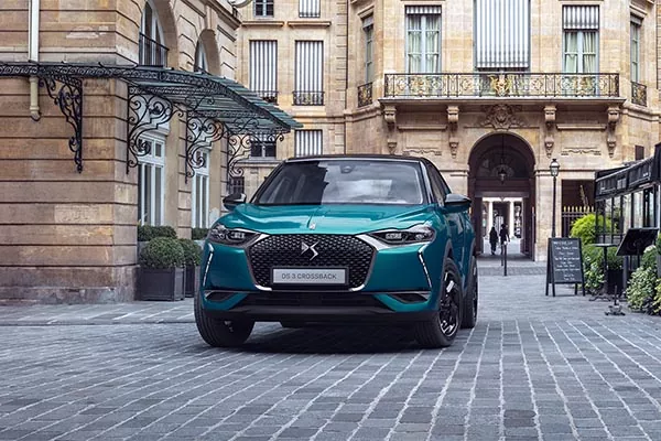 Ein tealfarbener DS 3 Crossback parkt auf einer Kopfsteinpflasterstraße vor eleganten, historischen Gebäuden mit großen Fenstern und verzierten Details.