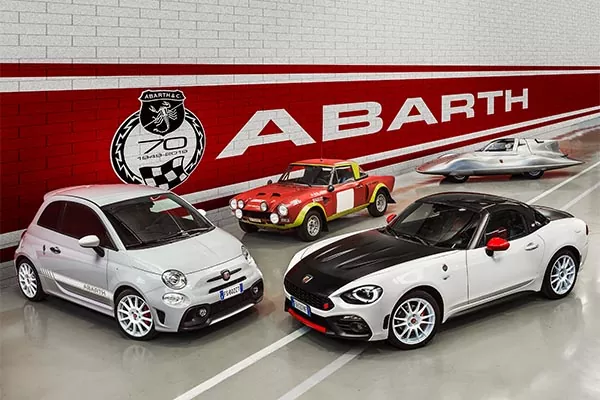 Vier Abarth-Autos - eine moderne Schräghecklimousine, ein modernes Cabriolet, ein klassisches Rallyeauto und ein Oldtimer-Rennwagen - parken vor einer rot-weißen Wand mit dem Abarth-Logo und einem Emblem zum 70-jährigen Jubiläum.