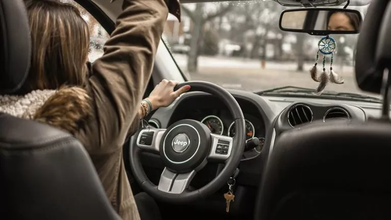Eine Person sitzt auf dem Fahrersitz eines Jeeps, hält mit einer Hand das Lenkrad und verstellt den Rückspiegel. Am Spiegel hängt ein Traumfänger, und die Autoschlüssel baumeln vom Zündschloss.