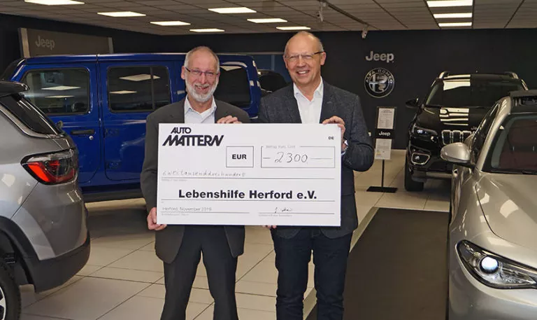 Zwei Männer in Anzügen stehen lächelnd in einem Autohaus und halten einen großen Spendenscheck über 2.300 Euro, ausgestellt auf die Lebenshilfe Herford e.V. Im Hintergrund sind Jeep-Fahrzeuge zu sehen.