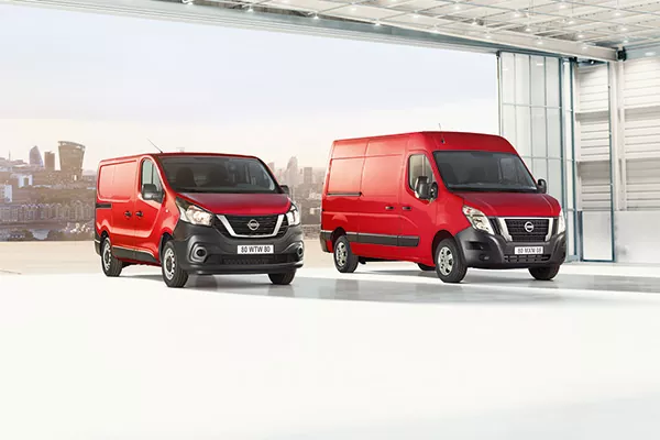 Zwei rote Nissan-Kleintransporter sind in einer hellen, modernen Garage mit großen Fenstern geparkt. Durch das offene Garagentor sind im Hintergrund städtische Gebäude zu sehen.