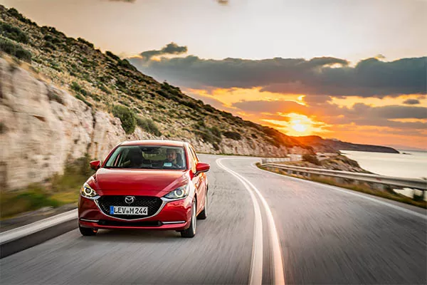 Ein rotes Auto fährt bei Sonnenuntergang eine kurvenreiche Küstenstraße entlang, mit felsigen Klippen auf der einen Seite und dem Meer in der Ferne unter einem dramatischen Himmel.