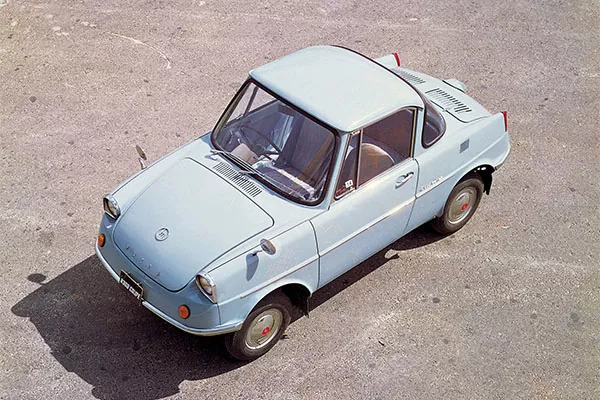 Ein kleiner, hellblauer Oldtimer mit zwei Türen und kompaktem Design ist auf einer gepflasterten Fläche geparkt, von oben gesehen. Der Wagen hat runde Scheinwerfer, eine kurze Motorhaube und rote Akzente an den Radnaben und Rückleuchten.