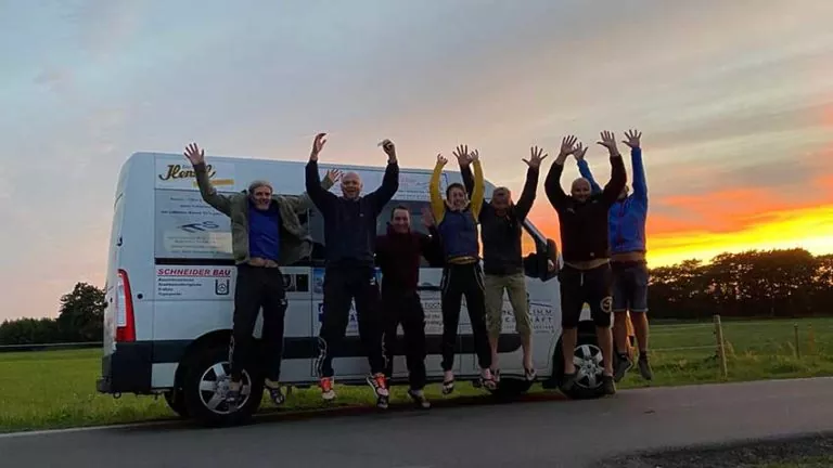 Sieben Personen springen mit erhobenen Armen vor einem weißen Lieferwagen in die Luft, der bei Sonnenuntergang auf einer Landstraße geparkt ist, mit Feldern und Bäumen im Hintergrund.