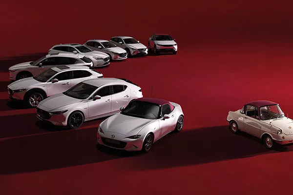Eine Reihe von neun Mazda-Fahrzeugen, meist weiß und modern, sind auf einem roten Boden angeordnet, wobei sich ein kleiner, weiß-roter Oldtimer von den neueren Modellen abhebt.