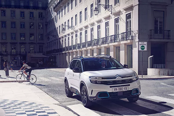 Ein weißer Citroën-Geländewagen parkt an einer sonnigen Straßenecke in der Nähe eines Schildes für eine Umweltzone, während in der Nähe eine Person Fahrrad fährt und im Hintergrund hohe Gebäude zu sehen sind.