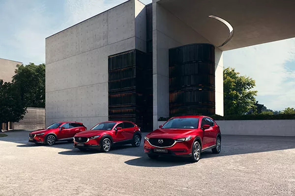 Drei rote Mazda-Geländewagen sind nebeneinander vor einem modernen Gebäude mit geometrischer Betonarchitektur geparkt, an einem sonnigen Tag mit Bäumen im Hintergrund.