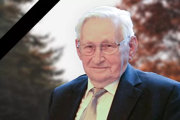 Ein älterer Mann mit weißem Haar und Brille, der Anzug und Krawatte trägt und leicht lächelt. Der Hintergrund besteht aus unscharfen Bäumen, mit einem schwarzen diagonalen Streifen in der oberen linken Ecke, was auf ein Gedenk- oder Tributbild hindeutet.