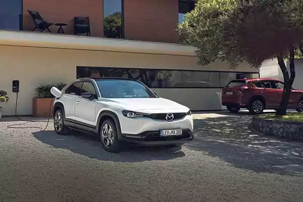 Ein weißer Mazda MX-30 Elektro-SUV ist vor einem modernen Haus geparkt und an eine Ladestation angeschlossen. Ein weiterer roter Geländewagen ist in der Nähe geparkt, während Bäume Schatten auf der Einfahrt spenden.