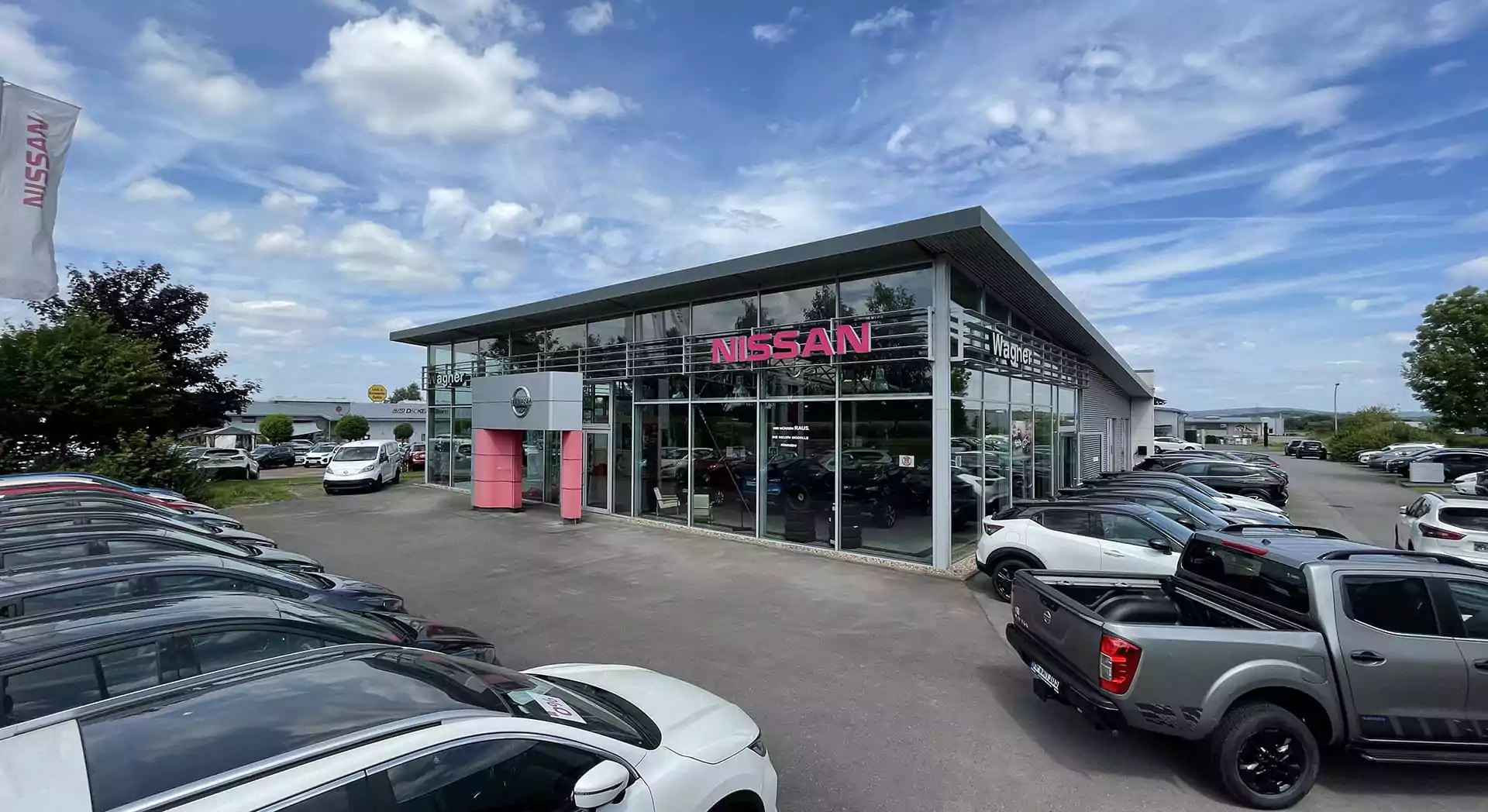 Ein modernes Autohaus Mattern Lage Nissan Autohaus mit großen Glasfenstern, mehreren geparkten Autos draußen und einem klaren Himmel mit vereinzelten Wolken darüber. Das Nissan-Logo und die Beschilderung sind auf dem Gebäude sichtbar.