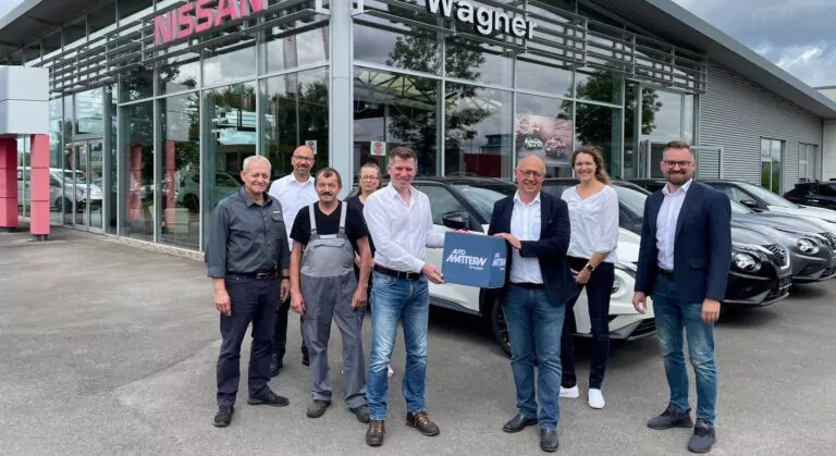 Sieben Personen stehen lächelnd vor einem Nissan-Autohaus der Wagner Lage Auto-Galerie. Ein Mann hält eine blaue Mappe mit der Aufschrift API MATTERN in der Hand, und neben dem gläsernen Gebäude sind mehrere Autos geparkt.