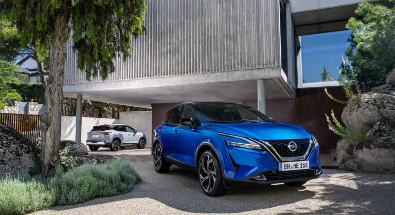 Ein blauer Nissan-Geländewagen ist auf einer Einfahrt vor einem modernen Haus geparkt, während im Hintergrund ein weißer Geländewagen unter einem von Bäumen und Felsen umgebenen freitragenden Bauwerk geparkt ist.