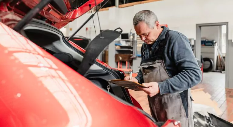 Ein Mechaniker im Overall inspiziert das Innere des Kofferraums eines roten Autos und schreibt Notizen auf ein Klemmbrett in einer gut beleuchteten Autowerkstatt, ideal für eine Stellenanzeige KFZ-Mechatroniker.