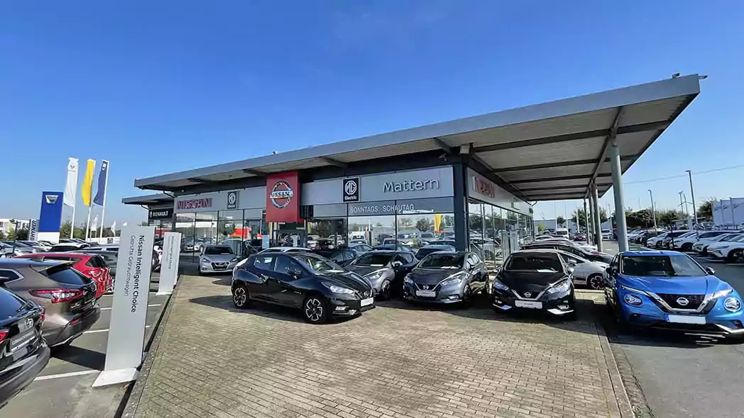 Ein modernes Nissan-Autohaus mit großen Glasfenstern zeigt mehrere Nissan-Fahrzeuge, die draußen auf einem gepflasterten Parkplatz unter blauem Himmel geparkt sind. Mehrere Autos und Schilder des Autohauses sind zu sehen.