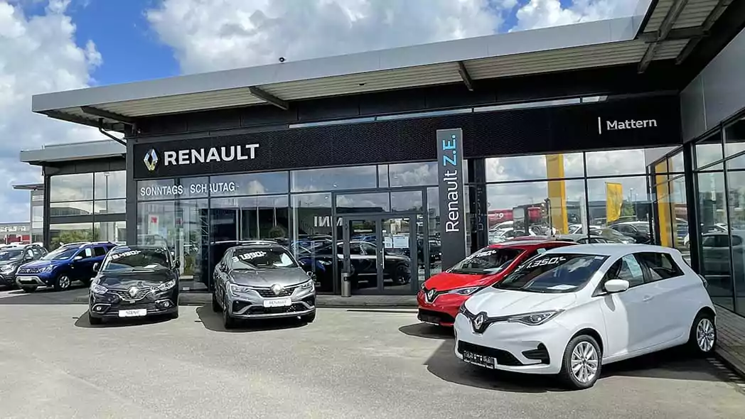 Vier Renault-Autos stehen vor einem modernen Renault-Autohaus mit großen Glasfenstern und einer auffälligen Beschilderung mit dem Renault-Logo und -Namen unter einem teilweise bewölkten Himmel.