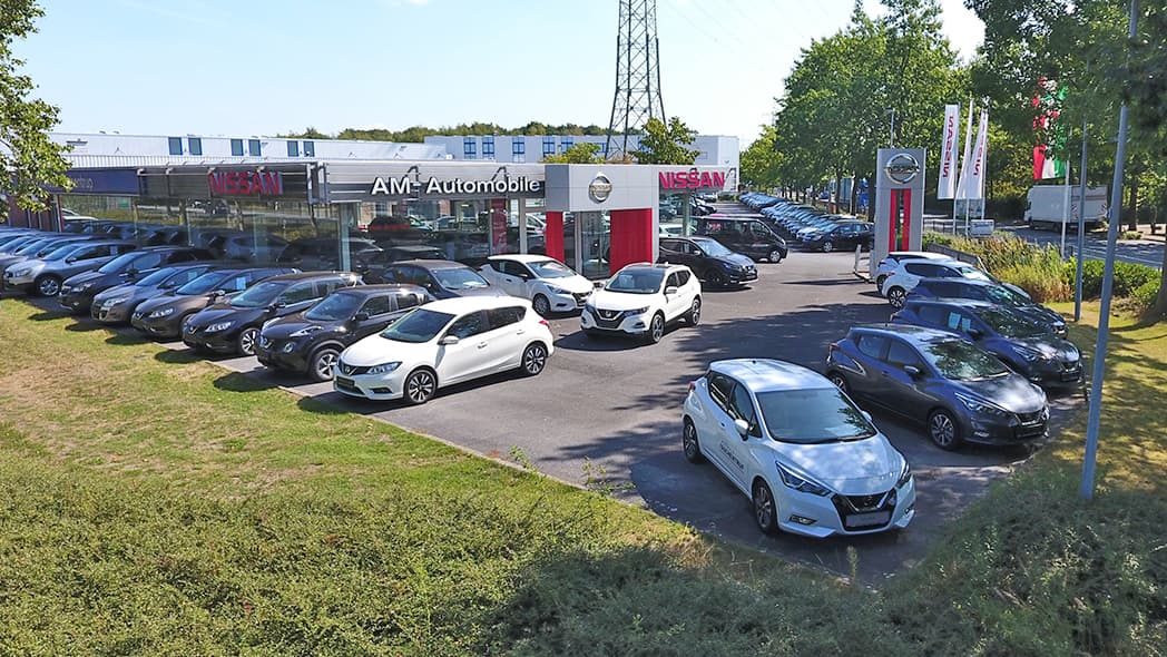 Luftaufnahme des Parkplatzes eines Nissan Autohauses mit mehreren geparkten Autos, hauptsächlich in Schwarz, Weiß und Grau, vor einem gläsernen Ausstellungsgebäude an einem sonnigen Tag.