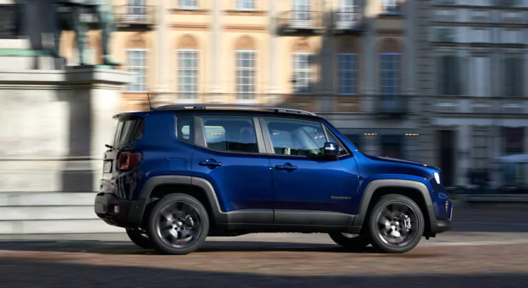 Ein blauer Jeep Renegade SUV fährt an einem verschwommenen Hintergrund historischer Gebäude mit großen Fenstern und Balkonen vorbei.