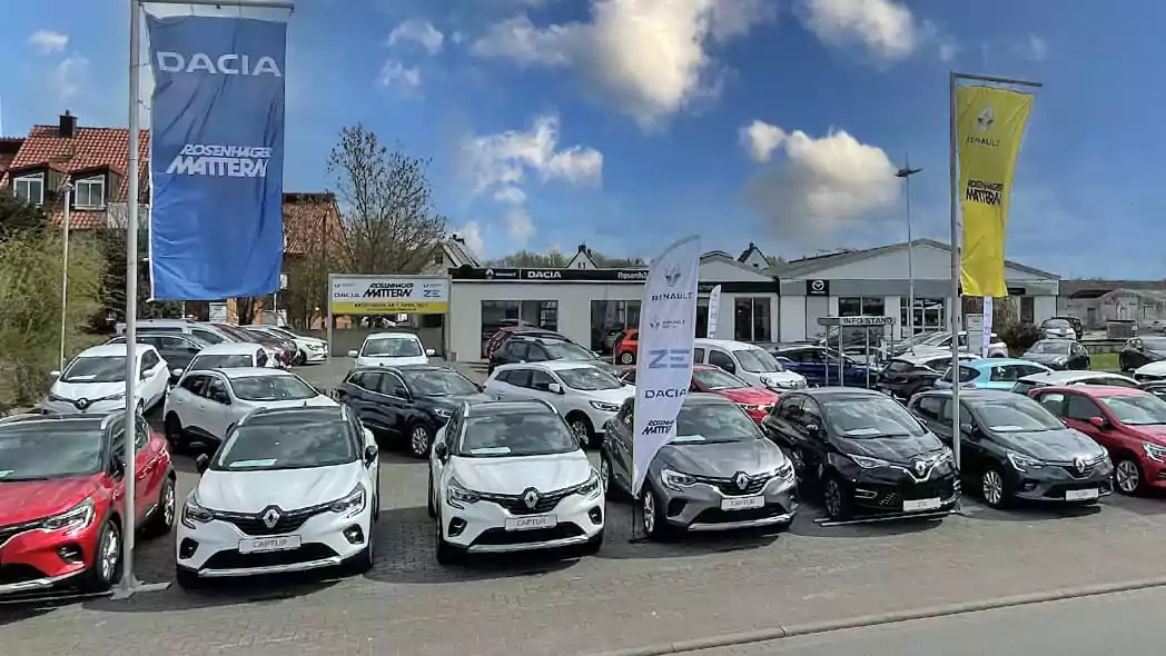 Ein Autohändlerparkplatz mit Reihen von neuen Dacia- und Renault-Fahrzeugen, die vor dem Renault Autohaus geparkt sind. Große blaue und gelbe Fahnen mit Dacia- und Renault-Logos sind zu sehen, während die Gebäude des Autohauses im Hintergrund unter einem teilweise bewölkten Himmel stehen.