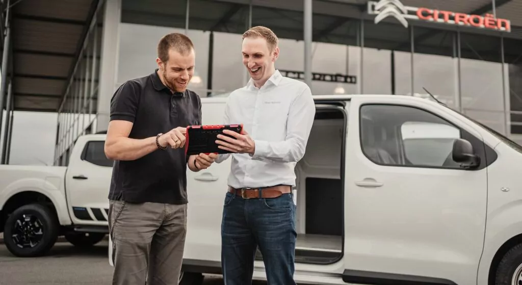 Zwei Männer stehen vor einem weißen Lieferwagen, schauen auf ein Tablet und lächeln. Im Hintergrund ist ein modernes Gebäude mit Citroën-Branding zu sehen.