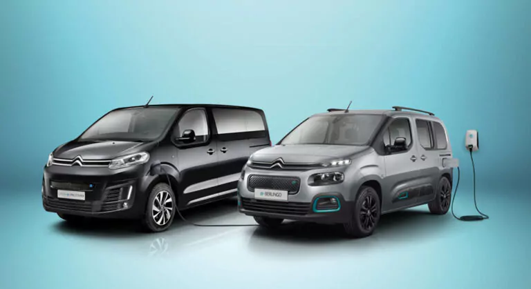 Zwei silberne Citroën-Elektrofahrzeuge sind nebeneinander vor einem blauen Hintergrund mit Farbverlauf geparkt; das Auto rechts ist an eine Wandladestation angeschlossen.