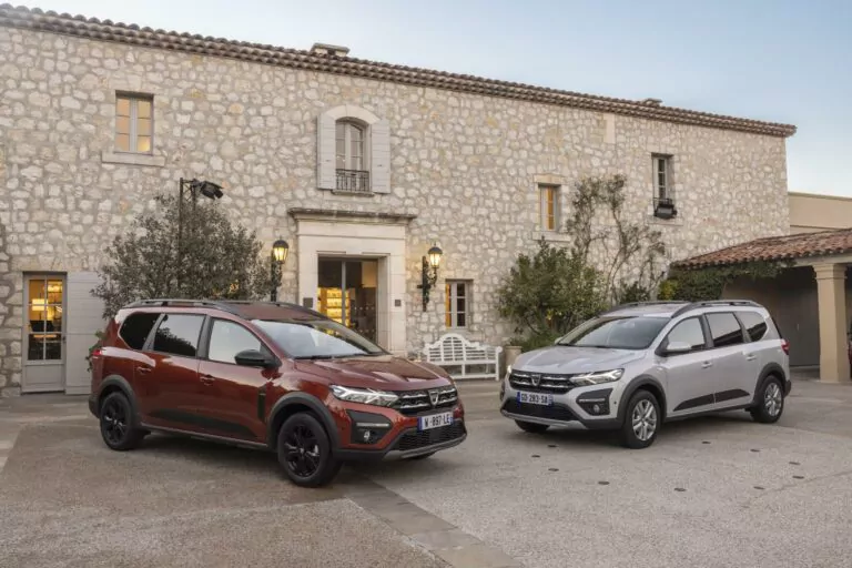 Zwei Dacia Jogger Kombi, ein roter und ein silberner, sind vor einem rustikalen Steingebäude mit mehreren Fenstern und einer Holztür geparkt, umgeben von Außenbeleuchtung und üppigem Grün.