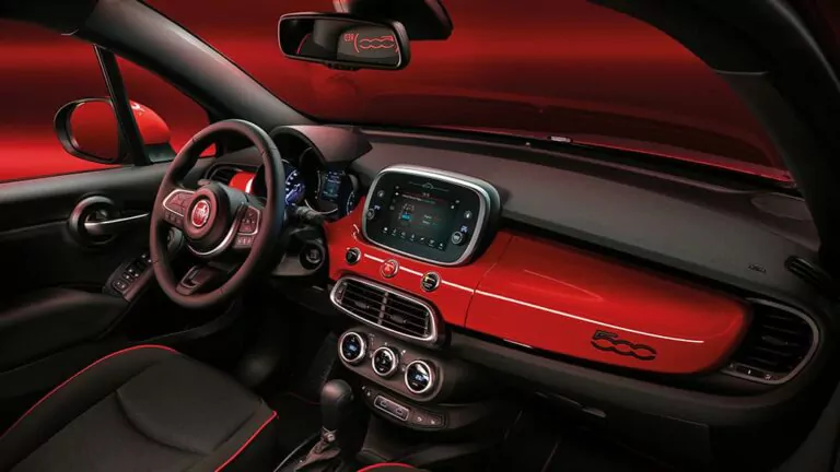 Innenraum eines modernen Fiat 500X Tipo Hybrid mit rot-schwarzem Armaturenbrett, Touchscreen-Display, runden Lüftungsdüsen und Fiat 500-Logo auf der Beifahrerseite. Der Innenraum verfügt über schwarze Sitze mit roten Akzenten und ein sportliches Lenkrad.