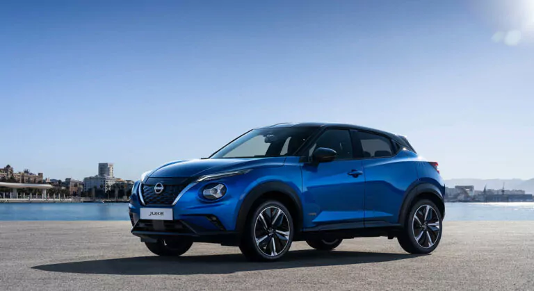 Ein blauer Nissan Juke, ein kompakter Geländewagen, parkt auf einer gepflasterten Fläche in der Nähe einer Uferpromenade, mit der Skyline der Stadt und einem klaren blauen Himmel im Hintergrund.