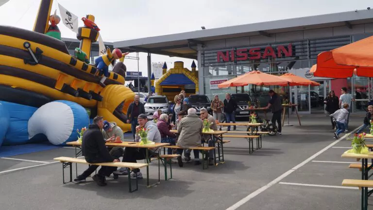 Während des Sommerfestes in Bielefeld sitzen die Menschen an Picknicktischen unter orangefarbenen Sonnenschirmen vor dem Nissan-Autohaus Auto Mattern, in der Nähe stehen aufblasbare Hüpfburgen in Form von Schiffen. Mehrere E-Autos und andere Autos sind geparkt, während Kinder und Erwachsene die Veranstaltung genießen.